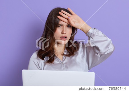 Tired student with headache posing isolated over lilac background and working with portable computer, looks at camera with exhausted facial expression, lady studying long hours and needs rest. Tired student with headache posing isolated over lilac background and working with portable computer, looks at camera with exhausted facial expression, lady studying long hours and needs rest. 76589544