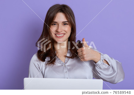 Attractive young brunette businesswoman showing thumbs up while working online indoor against lilac background, being in good news, dark haired lady with pleasant appearance online work. Attractive young brunette businesswoman showing thumbs up while working online indoor against lilac background, being in good news, dark haired lady with pleasant appearance online work. 76589548