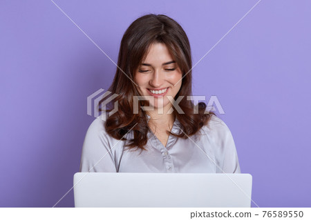Front view of attractive lady working on laptop and smiling, looks at screen, wearing elegant blouse, having pleasant news about work or study, female with portable computer against lilac wall. Front view of attractive lady working on laptop and smiling, looks at screen, wearing elegant blouse, having pleasant news about work or study, female with portable computer against lilac wall. 76589550