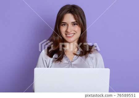 Portrait of adorable smiling woman holding white laptop isolated over lilac background, lady with good mood working online or studying, has friendly expression, girl with notebook indoor. Portrait of adorable smiling woman holding white laptop isolated over lilac background, lady with good mood working online or studying, has friendly expression, girl with notebook indoor. 76589551