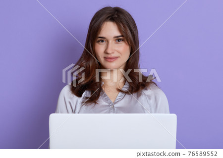 Beautiful caucasian young woman with laptop posing isolated over lilac background, looks at camera and smiling, wearing blouse, has dark hair, student or freelancer with portable computer. 76589552