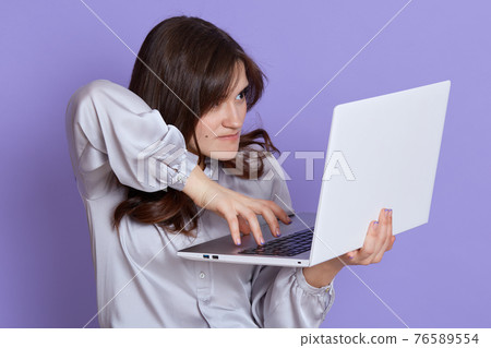 Angry executive elegant job woman typing on laptop, mad female working online, has lots work, looking at device screen with annoyed expression, wearing elegant blouse against lilac wall. 76589554
