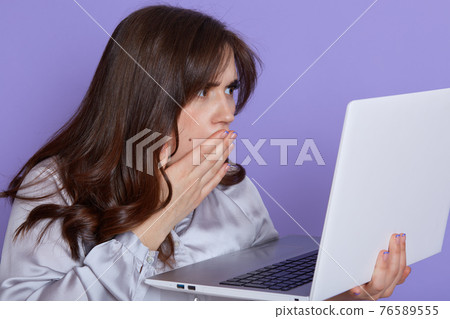 Amazed emotional woman with dark hair, looking at laptop screen with shocked expression, covering her opened mouth, isolated over lilac background, sees something bad, has trouble with work. Amazed emotional woman with dark hair, looking at laptop screen with shocked expression, covering her opened mouth, isolated over lilac background, sees something bad, has trouble with work. 76589555