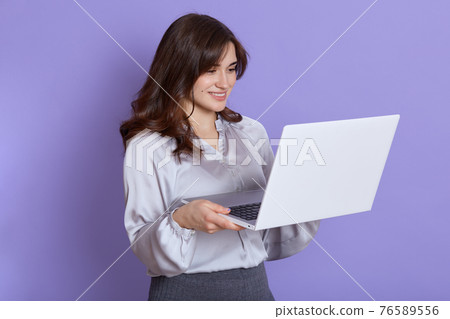 European young brunette woman girl in elegant white blouse and skirt posing isolated over lilac background, looking at screen with smile, winsome lady working online. European young brunette woman girl in elegant white blouse and skirt posing isolated over lilac background, looking at screen with smile, winsome lady working online. 76589556