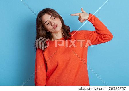 Horizontal shot of relaxed woman tilts head and shoots in temple, pretends killing herself, makes finger gun gesture, has tired expression, wears orange jumper, poses over blue wall. 76589767