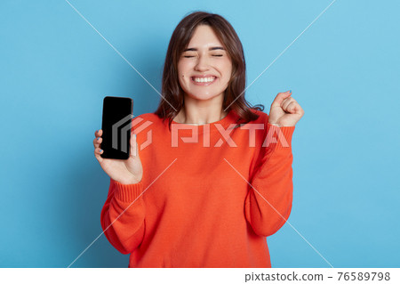 Overjoyed attractive woman keeps fist clenched, being happy to get good news, gets notification on cellphone, wears orange sweater, stands with closed eyes indoor against blue wall, feels amused. Overjoyed attractive woman keeps fist clenched, being happy to get good news, gets notification on cellphone, wears orange sweater, stands with closed eyes indoor against blue wall, feels amused. 76589798