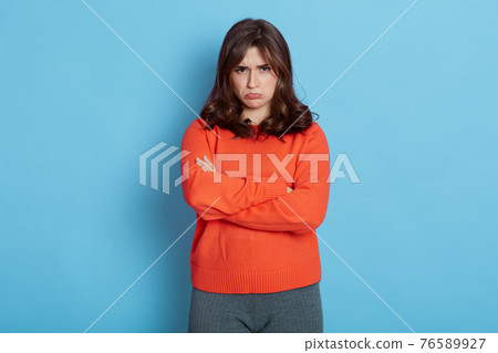 Upset young girl standing isolated over blue background with folded hands, looking at camera with pout lips, being very sad, expressing negative emotions, wearing orange sweater and leggins. Upset young girl standing isolated over blue background with folded hands, looking at camera with pout lips, being very sad, expressing negative emotions, wearing orange sweater and leggins. 76589927