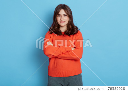 Smiling young brunette woman girl in sweater posing isolated blue background, alluring lady holding hands crossed, looks at camera with satisfied and happy expression. 76589928