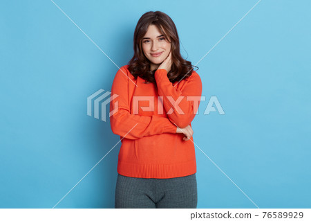 Cute brunette girl wearing casual sweater and grey leggins, looking at camera with shy charming smile and holding her neck, touched by nice compliment, expressing good feelings. Cute brunette girl wearing casual sweater and grey leggins, looking at camera with shy charming smile and holding her neck, touched by nice compliment, expressing good feelings. 76589929