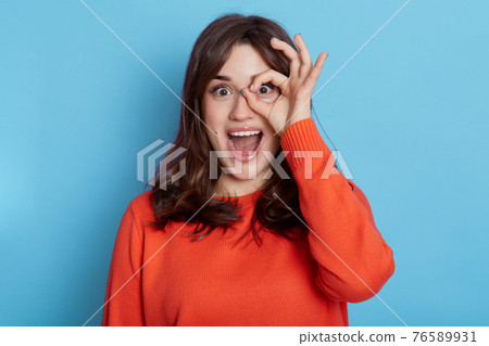 Appealing young woman making ok sign and covers her eye, shows tongue, expresses excitement, model poses against blue background, has good mood, girl with dark hair looks at camera with big eyes. Appealing young woman making ok sign and covers her eye, shows tongue, expresses excitement, model poses against blue background, has good mood, girl with dark hair looks at camera with big eyes. 76589931
