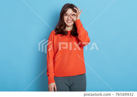 Funny girl showing gesturing ok sign with her hand like glasses and looking at camera with happy smile, appealing lady in casual jumper posing indoor, brunette female having fun. Funny girl showing gesturing ok sign with her hand like glasses and looking at camera with happy smile, appealing lady in casual jumper posing indoor, brunette female having fun. 76589932