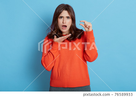 Surprised beautiful European female stares with bugged eyes and widely opened mouth, makes tiny gesture, posing against blue background., shows something too small, lady wearing casual attire. Surprised beautiful European female stares with bugged eyes and widely opened mouth, makes tiny gesture, posing against blue background., shows something too small, lady wearing casual attire. 76589933