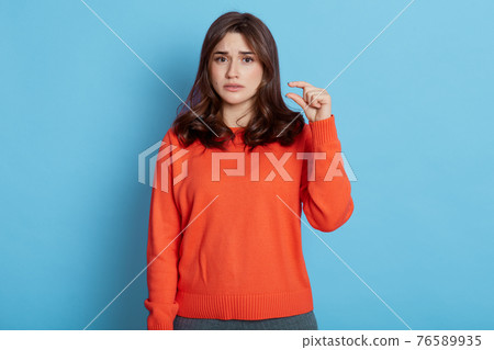 Sad beautiful lady shows tiny thing, makes small gesture, feels displeasure, dressed in casual clothes, stands against blue background, lady with unhappy look. Sad beautiful lady shows tiny thing, makes small gesture, feels displeasure, dressed in casual clothes, stands against blue background, lady with unhappy look. 76589935