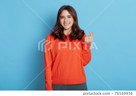 Cheerful young woman makes korean like sign, demonstrates her liking, expressing love,shows heart gesture with fingers, feels happy, wears orange jumper, dark haired girl isolated over blue background Cheerful young woman makes korean like sign, demonstrates her liking, expressing love,shows heart gesture with fingers, feels happy, wears orange jumper, dark haired girl isolated over blue background 76589936