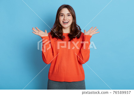 Happy pleased European woman keeps hands spreading aside, focused at camera, notices desirable things, laughs, dressed in orange sweater, poses against blue background. 76589937