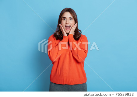 Portrait of amazed astonished beautiful female with surprised expression, poses against blue background, keeping hands on cheeks, poses with widely opened mouth, wearing orange sweater. Portrait of amazed astonished beautiful female with surprised expression, poses against blue background, keeping hands on cheeks, poses with widely opened mouth, wearing orange sweater. 76589938