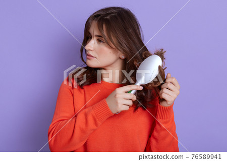 Sad young cute girl combing damaged hair, standing isolated on a pure background, looking away, combs tangled hair, wearing casual orange jumper, dark haired female. 76589941