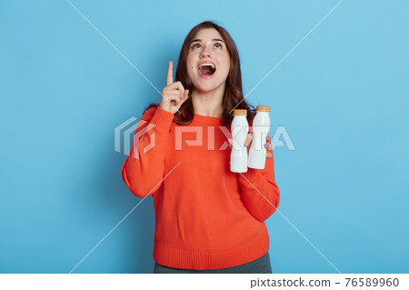 Young dark haired woman holding two bottles of milk, has idea or question pointing finger with happy and exited face, looking up, lady wearing casual shirt, keeping healthy diet. 76589960