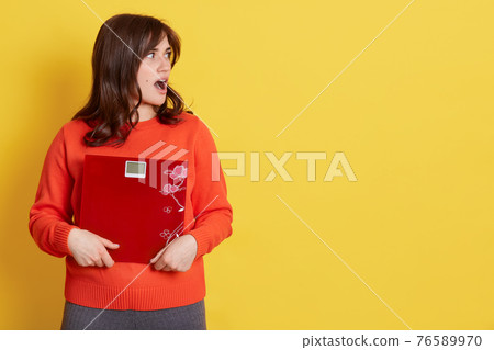 Brunette athletic woman holding scale and expressing surprise on face, looking aside with opened mouth, model posing isolated over yellow background, copy space for advertisement. Brunette athletic woman holding scale and expressing surprise on face, looking aside with opened mouth, model posing isolated over yellow background, copy space for advertisement. 76589970