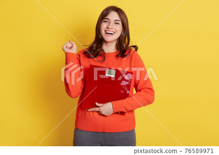 Winsome lady with floor scales in hands posing isolated over yellow background, clenching fist, being glad to slim down, finely loosing weight, girl wearing casual attire. Winsome lady with floor scales in hands posing isolated over yellow background, clenching fist, being glad to slim down, finely loosing weight, girl wearing casual attire. 76589971