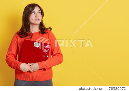 Diet and weight, young frustrated woman, looking aside with pensive facial expression, feeling unhealthy, embracing mechanical scales, wearing orange sweater, poses isolated over yellow background. Diet and weight, young frustrated woman, looking aside with pensive facial expression, feeling unhealthy, embracing mechanical scales, wearing orange sweater, poses isolated over yellow background. 76589972