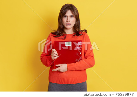 Frustrated woman with scale posing isolated over yellow background, lady being very upset and sad, female controlling her weight, has bad result, dresses casual clothing. 76589977