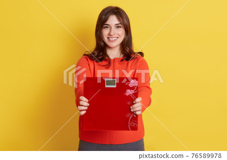 ult of loosing weight. Young attractive woman holding red floor scale in hands, looking directly nat camera, having joy facial expression, being very glad to have great resy ult of loosing weight. Young attractive woman holding red floor scale in hands, looking directly nat camera, having joy facial expression, being very glad to have great resy 76589978