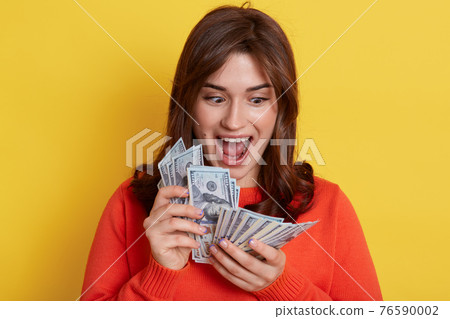Cheerful attractive young smiling woman holding cash, dreaming about its spending, keeps mouth widely opened, stands isolated over yellow background, lady with her salary. Cheerful attractive young smiling woman holding cash, dreaming about its spending, keeps mouth widely opened, stands isolated over yellow background, lady with her salary. 76590002