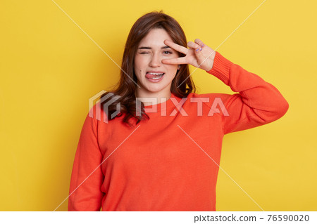 European dark haired lady with funny expression, posing isolated over yellow background, showing tongue, frowning face, winking one eye and cover other one with v sign, victory gesture. 76590020