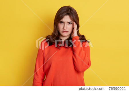 Distressed dark haired Caucasian woman touches temples and suffers migraine, has painful headache, frowns face, needs painkillers, wears in casual orange jumper, isolated over yellow background. Distressed dark haired Caucasian woman touches temples and suffers migraine, has painful headache, frowns face, needs painkillers, wears in casual orange jumper, isolated over yellow background. 76590021