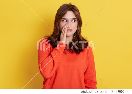 Mysterious dark haired woman keeps palm near mouth, looks aside and tells secret or private information, lady wearing in orange jumper gossips about something, stands indoor against yellow wall. Mysterious dark haired woman keeps palm near mouth, looks aside and tells secret or private information, lady wearing in orange jumper gossips about something, stands indoor against yellow wall. 76590024