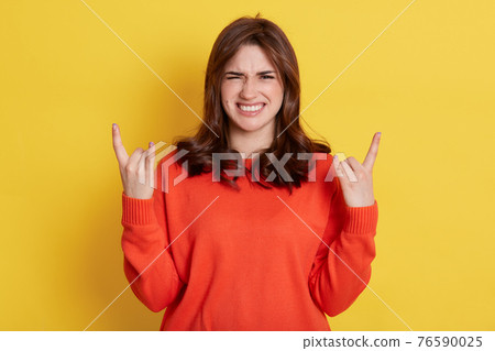 Cheerful emotional dark haired female makes rock n roll gesture and yells loudly, excited lady with happy frowning face, posing isolated over yellow background. 76590025