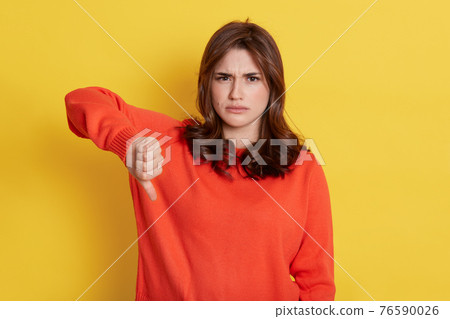 Teenager girl wearing orange sweater showing thumb down sign with sad facial expression, looks at camera with upset look, standing isolated over yellow background. Teenager girl wearing orange sweater showing thumb down sign with sad facial expression, looks at camera with upset look, standing isolated over yellow background. 76590026