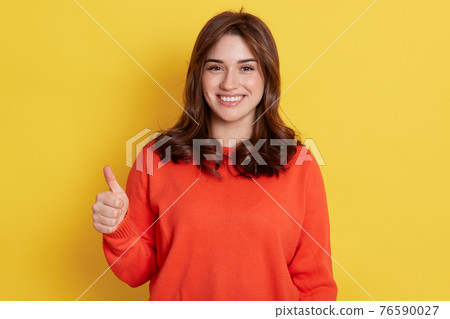 European happy young woman wearing orange clothes, holds finger up, shows like with thumb up isolated over yellow background, girl expressing positive, has satisfied look. 76590027