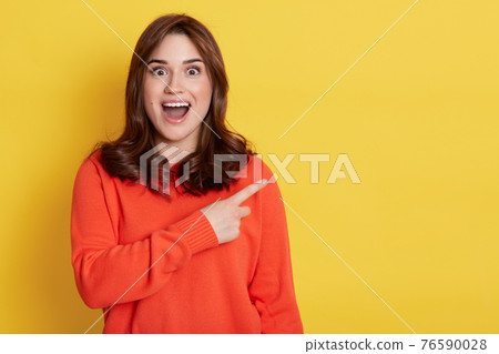 Young European excited young brunette woman girl in orange sweater, posing isolated on yellow background, beautiful lady with opened mouth pointing index finger aside. 76590028