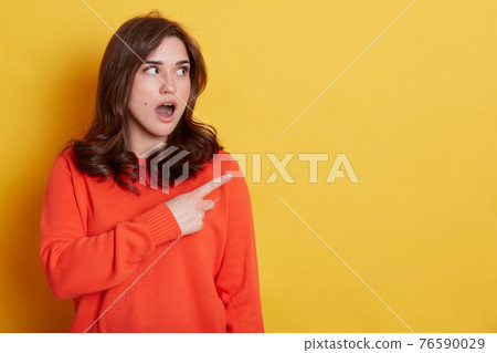 Portrait of shocked caucasian female with pleasant appearance indicates aside at blank yellow wall, has surprised and astonished expression, demonstrates something amazing. 76590029