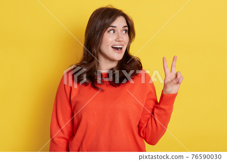 Portrait of joyful smiling pretty girl wearing orange jumper, showing v sign with fingers and looking aside with excited facial expression, shows peace or victory gesture, rejoicing success. Portrait of joyful smiling pretty girl wearing orange jumper, showing v sign with fingers and looking aside with excited facial expression, shows peace or victory gesture, rejoicing success. 76590030