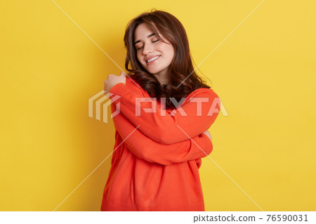 Lovely happy European woman daydreams, embraces herself with both hands, recalls romantic event, feels comfort, has broad smile and stands with closed eyes, wears orange casual sweater. Lovely happy European woman daydreams, embraces herself with both hands, recalls romantic event, feels comfort, has broad smile and stands with closed eyes, wears orange casual sweater. 76590031