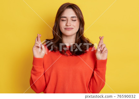 Pretty superstitious woman crosses fingers, prays for better, looks calm and relaxing, closes eyes, wears orange jumper, poses indoor over bright yellow background, anticipates important results. Pretty superstitious woman crosses fingers, prays for better, looks calm and relaxing, closes eyes, wears orange jumper, poses indoor over bright yellow background, anticipates important results. 76590032