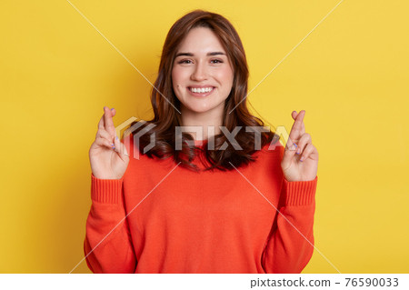 Happy dark haired female believing dreams come true. Hopeful woman feels lucky, crosses fingers, awaits for miracle, having toothy smile, wears casual sweater, poses against yellow wall. 76590033