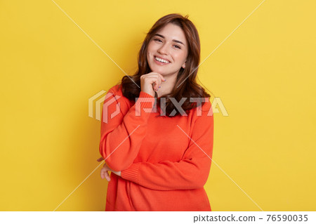 European charming girl with dark hair, holding fist under chin and smiling broadly, looking directly at camera, posing isolated over yellow background, expressing happiness. 76590035