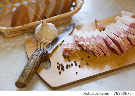 Horizontal indoor picture of wooden cutting board with salo, knife, peppercorn and garlic on it, delicious tasty food, traditional Ukrainian appetizer, composition. People and traditions concept. 76590675