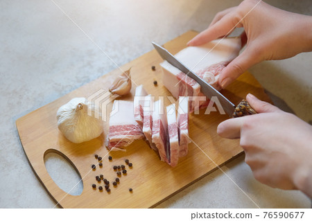 Faceless portrait of woman cutting lard with layers of meat on wooden cutting board among some garlic, peppercorns on white table, females's hands cuts fat, lady preparing snack for her husband. Faceless portrait of woman cutting lard with layers of meat on wooden cutting board among some garlic, peppercorns on white table, females's hands cuts fat, lady preparing snack for her husband. 76590677