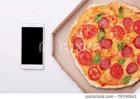 Pizza in box and smartphone with blank screen on white table, top view of delicious pepperoni isolated over light background. Modern mobile phone near box with pizza. Copy space for advertisment. Pizza in box and smartphone with blank screen on white table, top view of delicious pepperoni isolated over light background. Modern mobile phone near box with pizza. Copy space for advertisment. 76590681