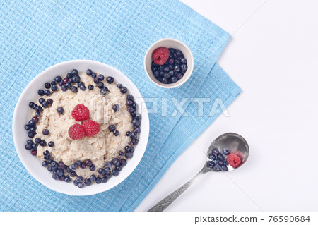 Photo of deep plate of oatmeal with strawberries and raspberries stand on delicate white wooden background on which lies blue cotton towel. Healthy eating, breakfast, healthy food and diet concept. 76590684