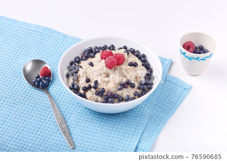 Top view of healthy delicious breakfast, oatmeal with blueberries and raspberries on white plate on blue table and spoon. Health care and healthy eating concept. Ready breakfast on white table surface Top view of healthy delicious breakfast, oatmeal with blueberries and raspberries on white plate on blue table and spoon. Health care and healthy eating concept. Ready breakfast on white table surface 76590685