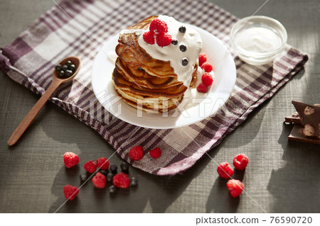 Top view of tasty pancakes with blueberries and raspberries on dark brown table, cup of tea or coffe, wooden spoon with fresh berry, pieces of chocholate. Delicious pancackes for family breakfast. Top view of tasty pancakes with blueberries and raspberries on dark brown table, cup of tea or coffe, wooden spoon with fresh berry, pieces of chocholate. Delicious pancackes for family breakfast. 76590720