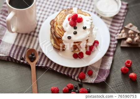 Pancackes with sour cream and tea on dark surface, top view of table decorated with muffins, raspberries, blueberries, spoon, chocolate, cup of coffe or tea, cotton towel. Tasty breakfast in morning. 76590753