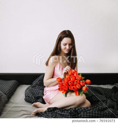 Young beautiful woman sitting in bed and holding a bouquet of red tulips Young beautiful woman sitting in bed and holding a bouquet of red tulips 76591073