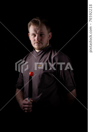 A chef in a uniform jacket shows off a professional chef's knife and a tomato 76592218